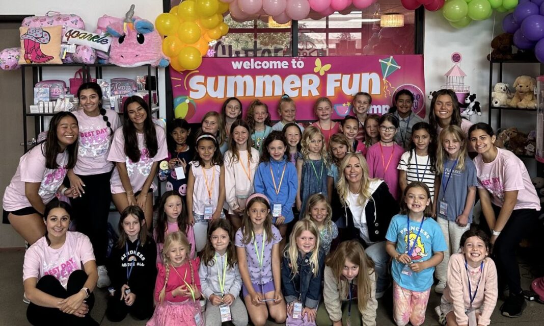 A group of smiling children and camp counselors pose at a summer camp in Rochester Hills, surrounded by colorful decorations, balloons, and fun-themed merchandise.
