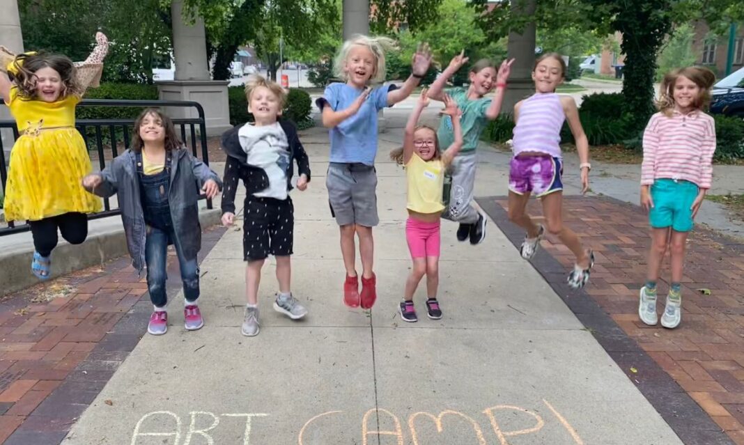 Excited kids jumping in the air at Art Explorers Summer Camp at Riverside Arts Center, with 'Art Camp!' written in colorful chalk on the sidewalk. A fun and creative summer camp experience for children.