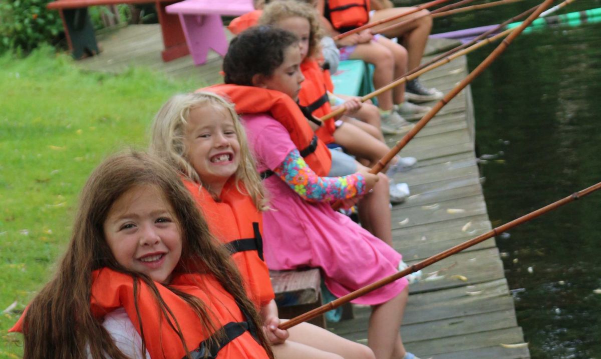 A group of smiling children wearing orange life jackets sit on a wooden dock at Willoway Day Camp, holding fishing poles over the water.