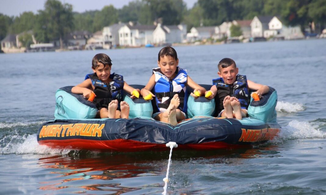 Three smiling boys wearing life jackets enjoy an exciting tubing ride on the lake at Willoway Day Camp, holding onto the inflatable raft as they glide across the water.