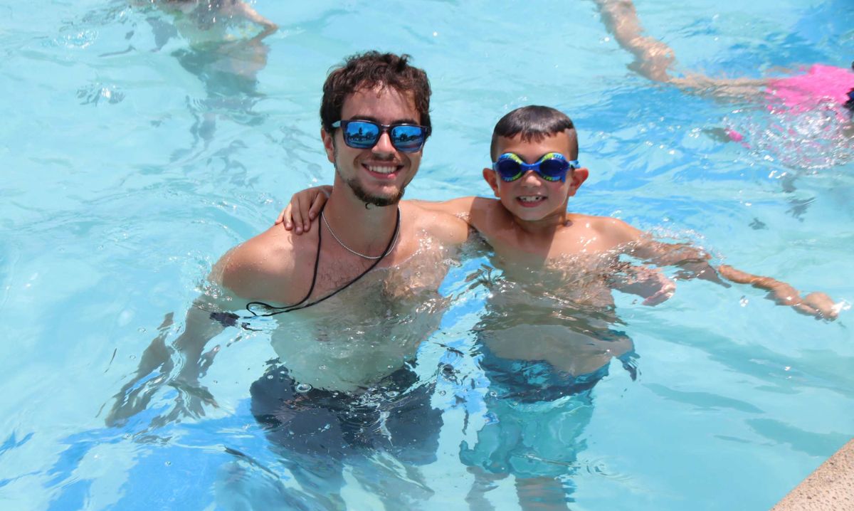 A smiling swimming instructor and a young camper wearing goggles pose together in the pool at Willoway Day Camp on a sunny day.