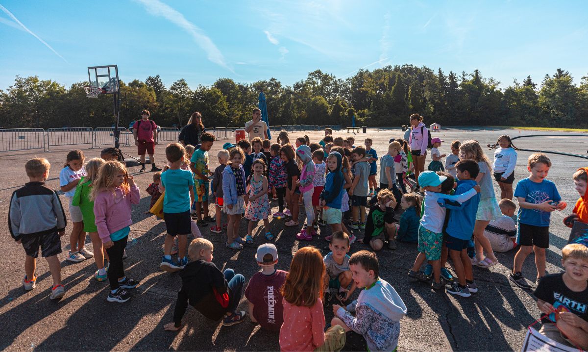 A large group of children gathers outdoors at Camp Mirage in Michigan for a summer camp activity. The bright, sunny setting highlights the excitement of outdoor games, socializing, and fun-filled experiences at this popular kids' camp.
