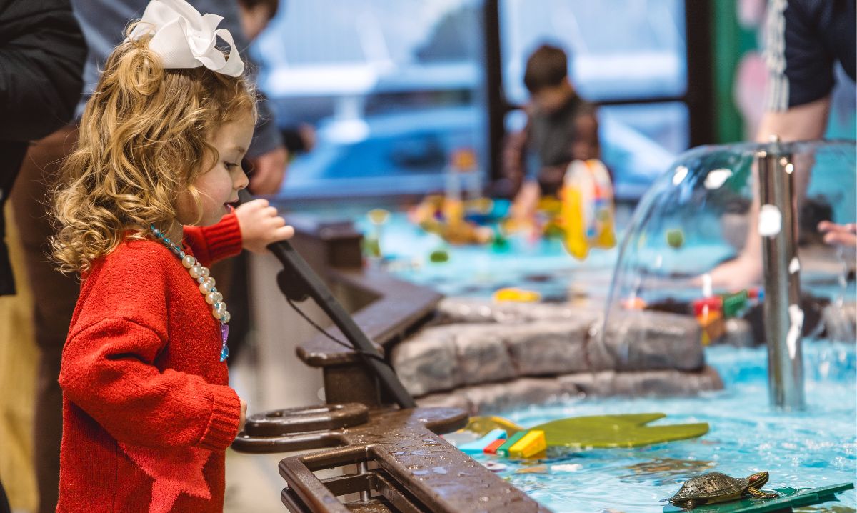A young girl in a red sweater exploring a hands-on water exhibit at a children's museum in Hocking Hills, Ohio, engaging in interactive learning and play.