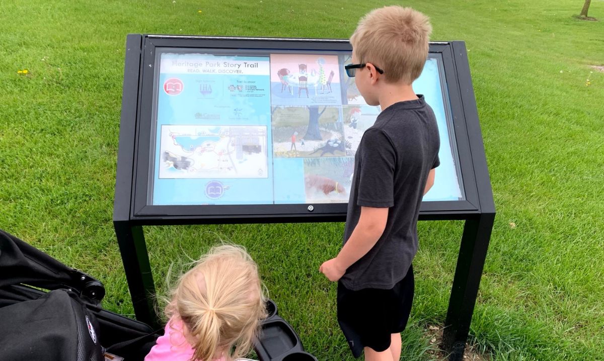 A young boy and a toddler explore an interactive story trail sign at Heritage Park, engaging with outdoor reading and learning activities.