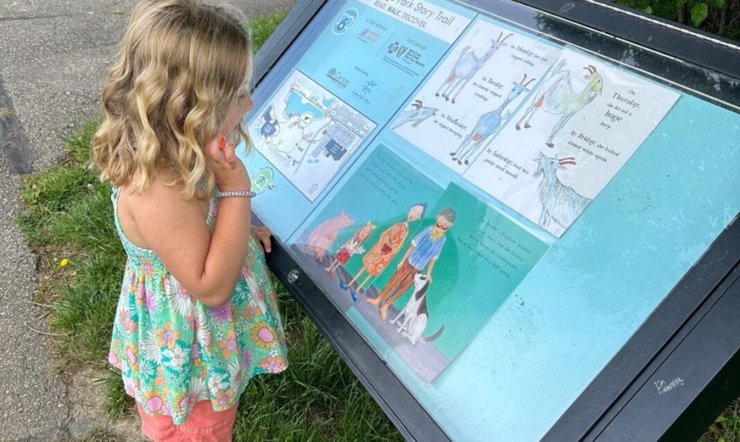 A young girl in a floral dress explores a story trail sign at Heritage Park, reading an illustrated children's story outdoors on a sunny day.
