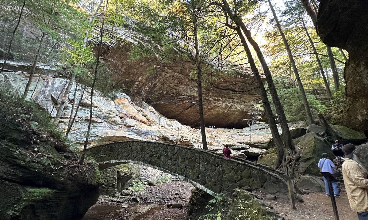 A scenic view of a stone bridge leading to a massive rock shelter at Hocking Hills, Ohio, surrounded by lush forest and towering cliffs, a popular hiking destination.