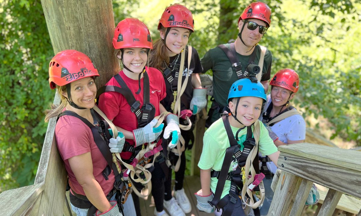 A smiling family dressed in safety harnesses and helmets, preparing for a ziplining adventure at Hocking Hills Zipline Adventures in Ohio, surrounded by lush green trees.