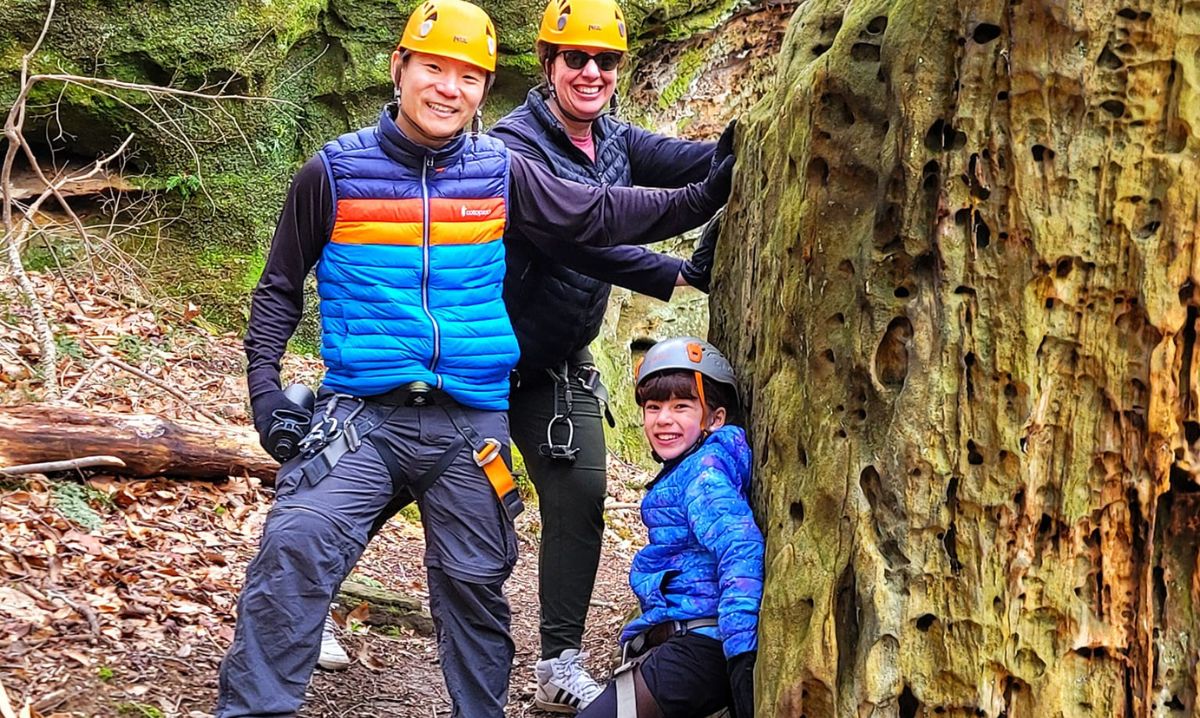 A family wearing helmets and harnesses enjoying a guided ziplining and hiking adventure in Hocking Hills, Ohio, surrounded by rugged rock formations and lush forest scenery.