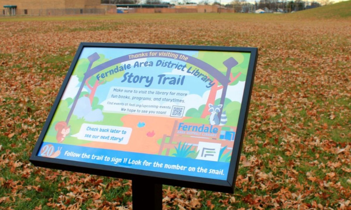 A story trail sign at Martin Road Park in Ferndale, Michigan, marking the end of the Ferndale Area District Library Story Trail. The colorful sign features a raccoon and squirrel illustration, library branding, and a message encouraging visitors to check back for new stories and visit the library for books and events. Fallen autumn leaves cover the grassy field in the background.