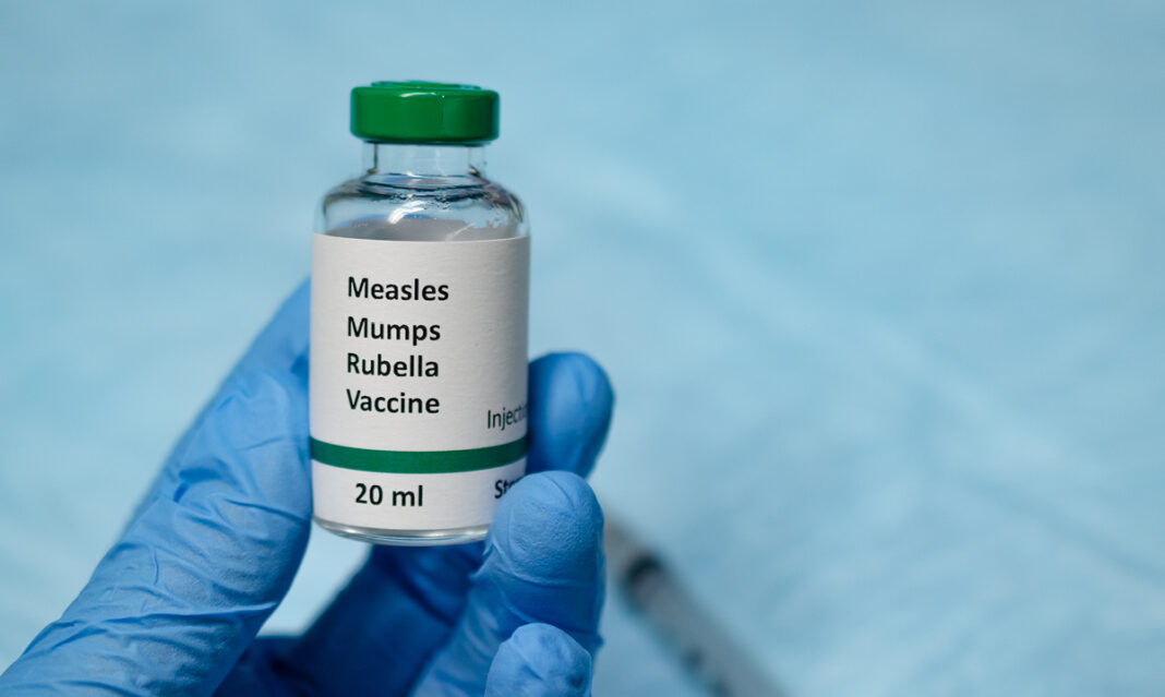 A healthcare professional wearing blue gloves holds a vial of the measles, mumps, and rubella (MMR) vaccine. Measles vaccine availability in Michigan is crucial for preventing outbreaks.