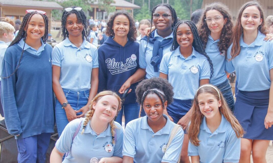 A group of smiling girls wearing Blue Lake Fine Arts Camp uniforms pose together outdoors with other campers in the background.