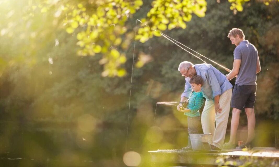 A multigenerational family enjoys a fishing trip in the United States, with a grandfather teaching his young grandson how to fish while the father looks on. The peaceful outdoor setting highlights the bonding experience and tradition of family fishing trips in the United States.