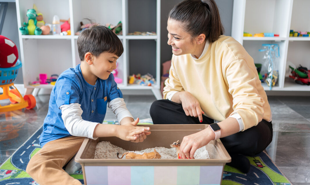 A young boy and a therapist engage in sand play therapy in a bright and colorful playroom, using toy animals to interact and communicate.