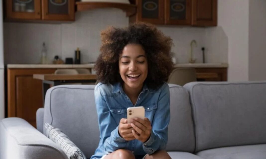 A young girl smiles while using a smartphone at home, raising the question of how young is too young for a smartphone. Parents often debate the right age for kids to have their own devices.