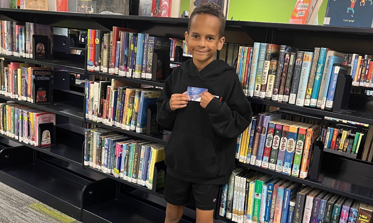 A young boy proudly holding his library card at Mount Clemens Public Library, standing in front of bookshelves filled with colorful children's books.