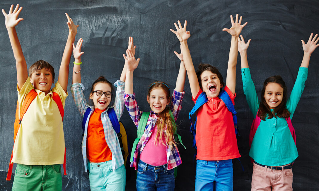 Group of smiling elementary school kids with backpacks raising their hands in front of a chalkboard, representing positive strategies for boosting school attendance.