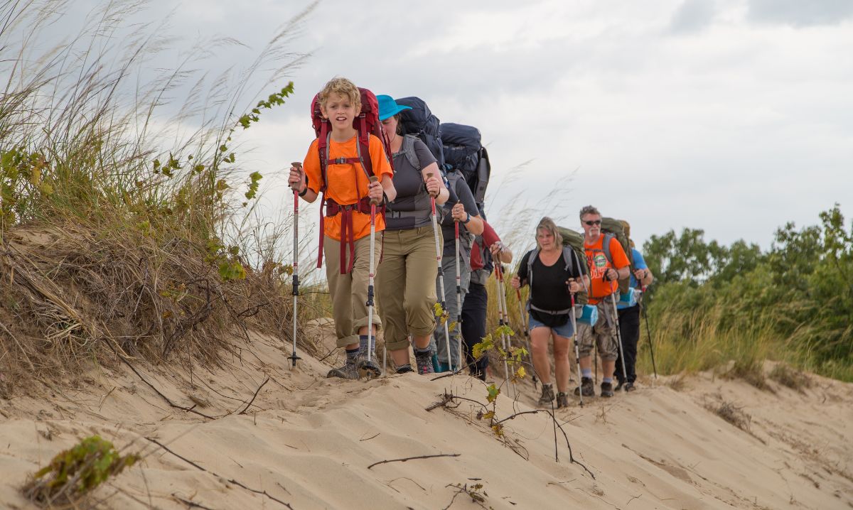 Multigenerational hiking group trekking through sandy trails and grassy dunes at Indiana Dunes National Park on a cloudy day.