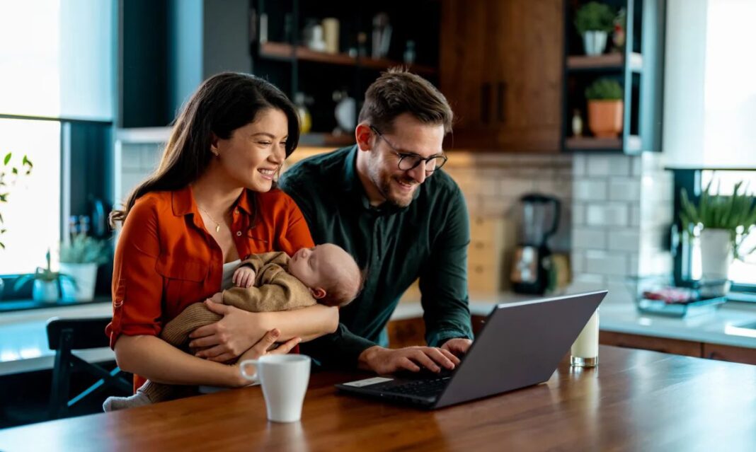 A young family sitting in their kitchen smiling at a laptop while holding a baby, symbolizing the beginning steps of how to create a spending plan together.
