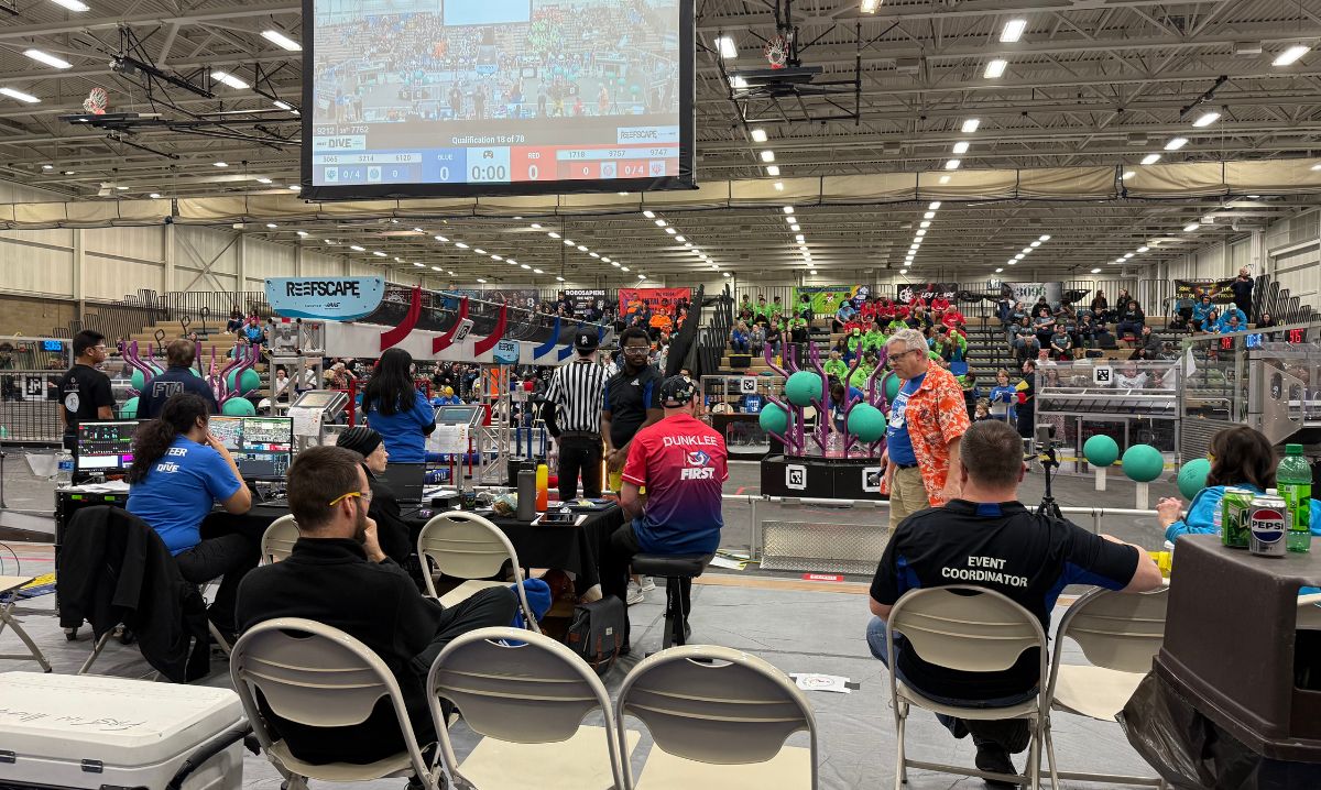 Event staff and volunteers manage scoring and operations at the MARZ FIRST Robotics competition, with colorful team sections and the playing field in the background.