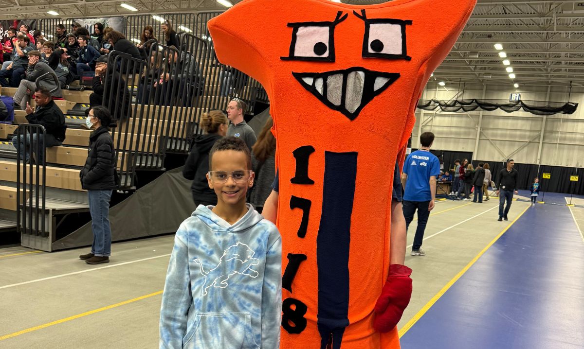 A young student poses with the Team 1718 mascot at the MARZ FIRST Robotics competition, with spectators seated in the background.