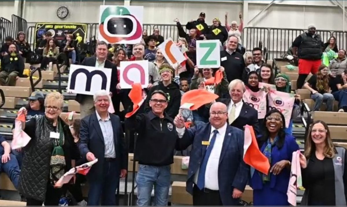 Community supporters and local leaders cheer on MARZ robotics team at a school event, holding signs and waving team flags in the bleachers