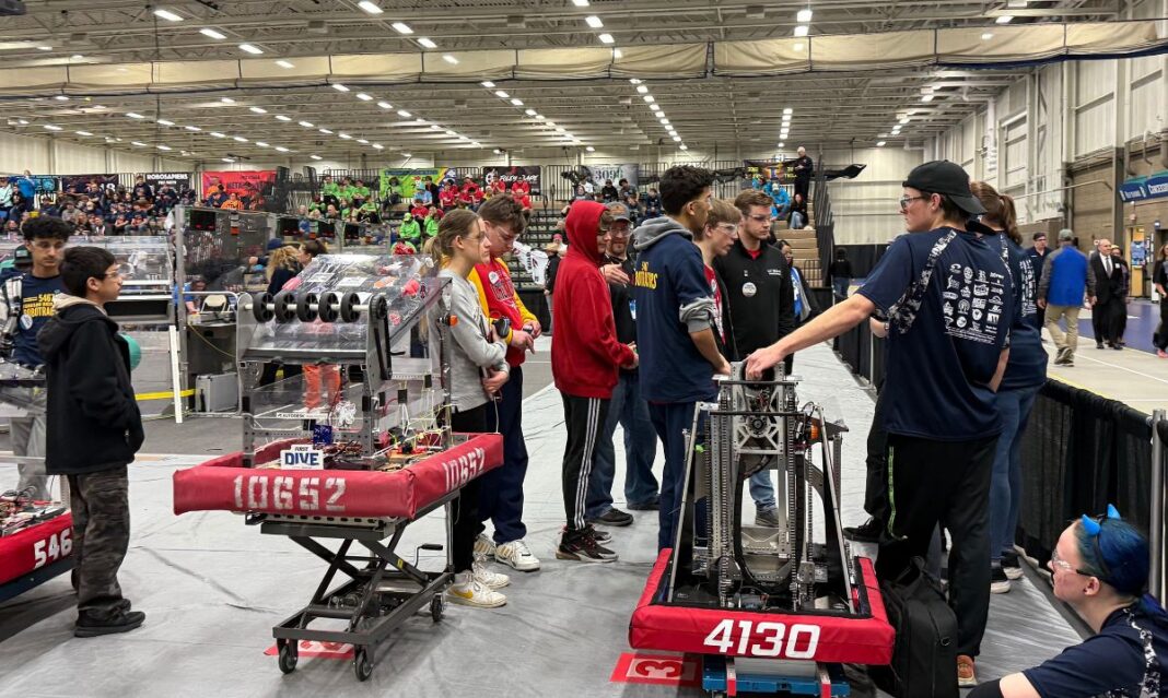 Students from multiple robotics teams discuss strategy and inspect their robots during a FIRST Robotics competition event in a packed gymnasium