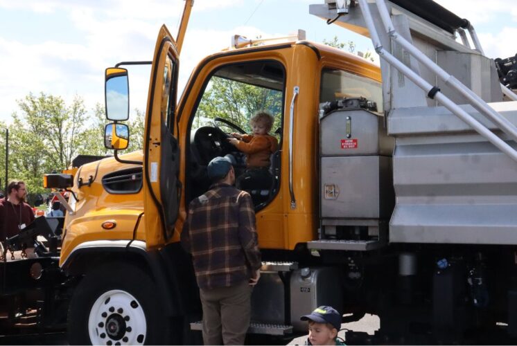 Child sitting in the driver’s seat of a yellow snow plow truck during a Touch-a-Truck community event in Metro Detroit.