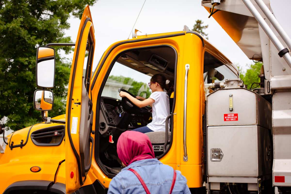 Young girl sitting in the driver’s seat of a large yellow utility truck at Truck-a-Palooza, a hands-on event hosted by Farmington Community Library for kids in Metro Detroit.