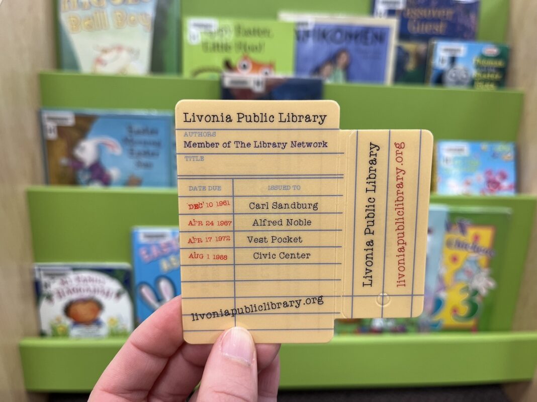A hand holding a Livonia Public Library card designed like a vintage checkout slip, with colorful children’s books on shelves in the background — how to get a library card in metro Detroit.