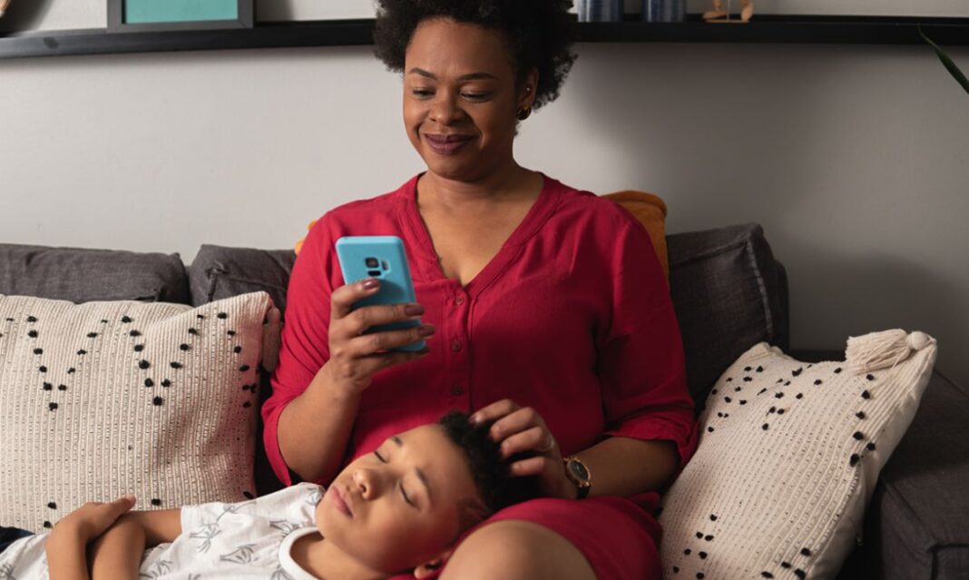 Parent using mental health app on smartphone while child rests, promoting emotional wellness with Oakland Community Health Network