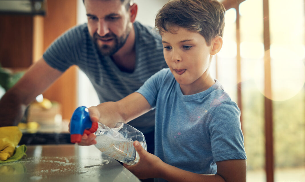 Young boy with autism spraying cleaner on a countertop while his dad supports and encourages him during a household chore