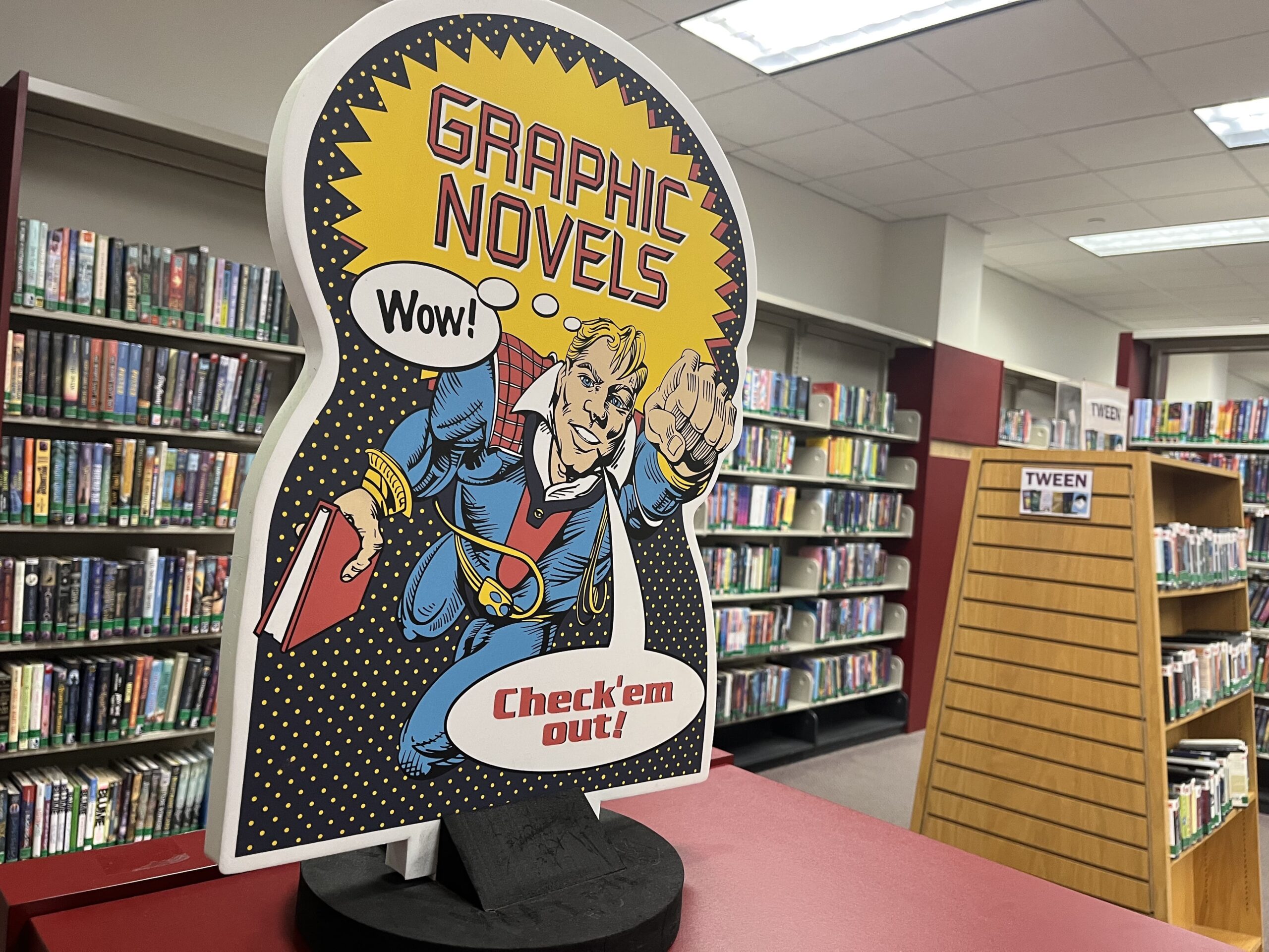 The graphic novel section of the Livonia Public Library, featuring a dynamic comic-style sign.
