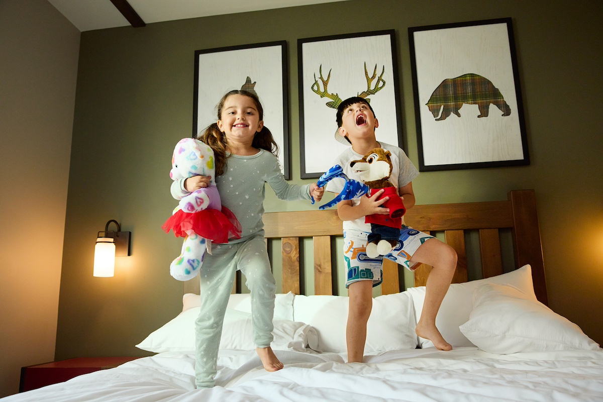 Two kids playing with stuffed animals and jumping on the bed in a guest room at Great Wolf Lodge Traverse City.