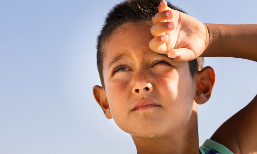 Close-up of a child squinting and shielding their eyes from the sun, showing flushed cheeks—a possible sign of heat rash in kids during hot summer days in Metro Detroit.