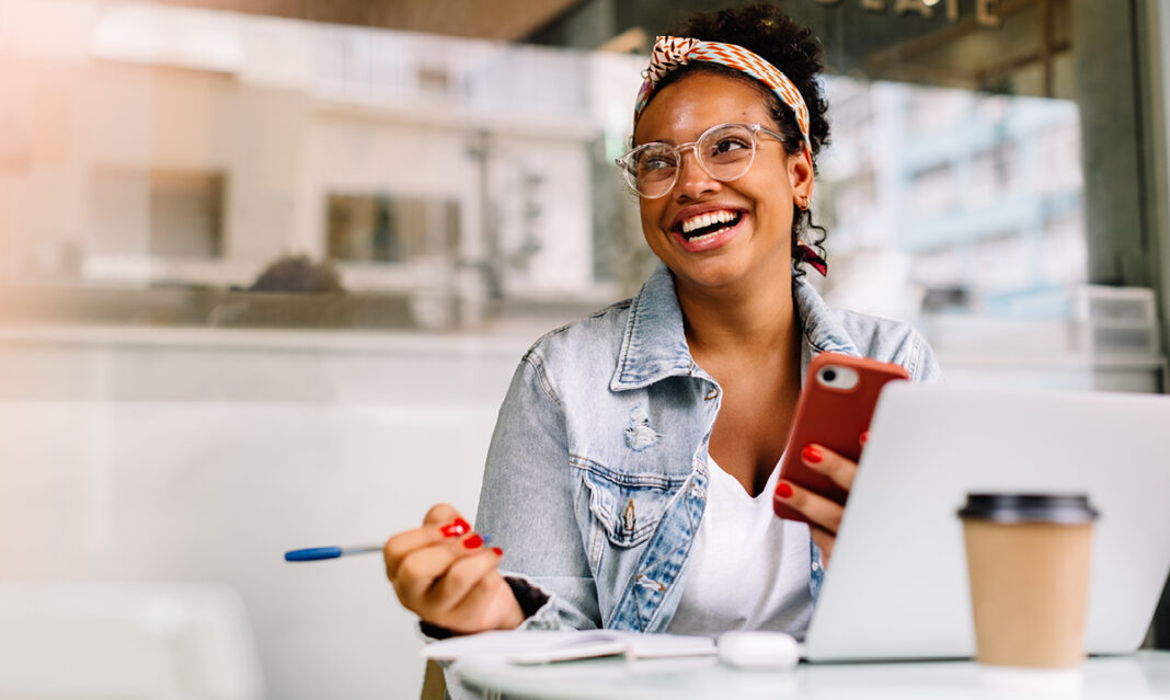 Smiling college student in a café using a smartphone and laptop, representing digital tools and resources for how to pay for college.