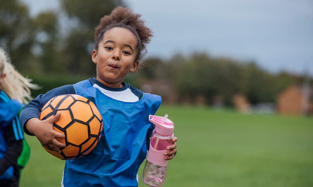 Young girl in a soccer jersey holding a ball and water bottle on a grassy field, representing youth sports, hydration, and nutrition for active kids in Metro Detroit.