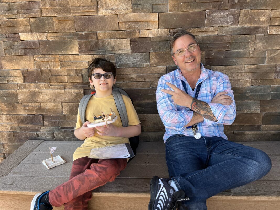 Teacher and student at Aim High School sitting on a bench outside, smiling and holding a class project, representing supportive special education accommodations.