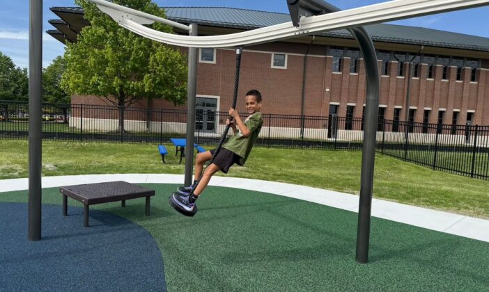 Boy rides accessible zip line at Clinton Township inclusive playground, featuring adaptive equipment and sensory-friendly surfaces for all abilities.