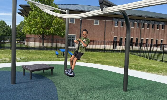 Boy glides along adaptive zip line at Clinton Township inclusive playground, designed with accessible play equipment and safe, rubberized surfaces.