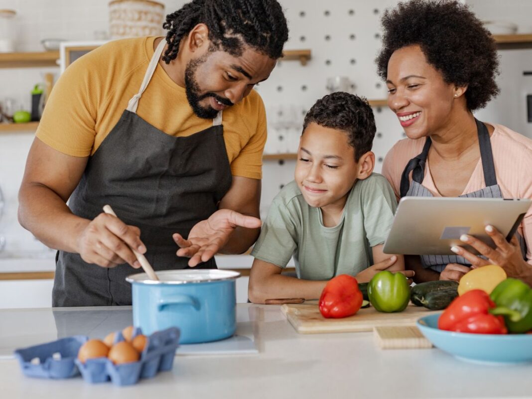 A smiling family of three cooks together in a modern kitchen—dad stirs a pot while mom follows a recipe on a digital tablet and their child looks on—illustrating the convenience and togetherness of following an AI meal plan.