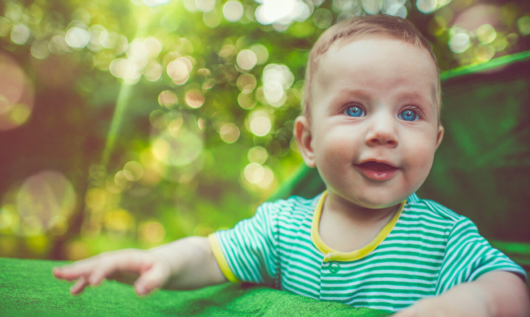 Smiling baby with blue eyes dressed in lightweight clothing enjoying shade outdoors, representing summer heat safety tips for babies in Metro Detroit.