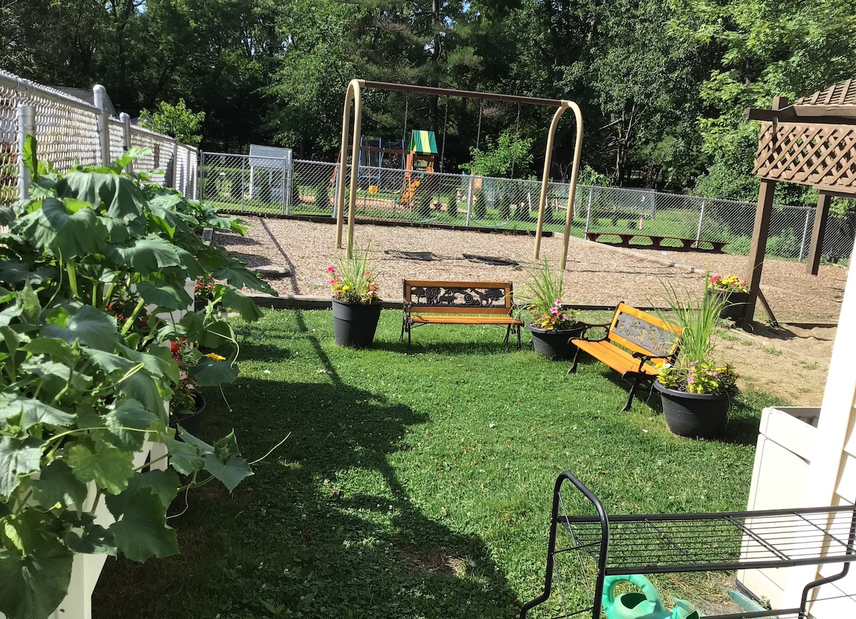 Outdoor playground and garden seating area at Novi Northville Montessori Center, featuring swing set, benches, flowers, and lush greenery for student exploration and outdoor learning.