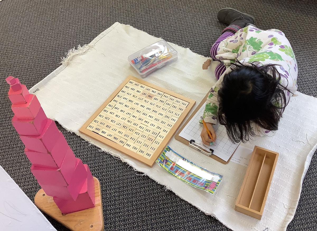 A young student at Novi Northville Montessori Center works on a math activity using the Montessori hundred board while lying on a classroom rug.