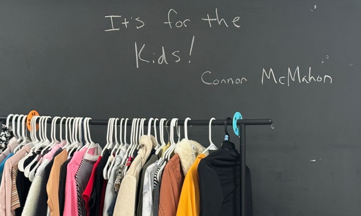 Clothing rack at the thrift store in Fraser High School with a chalkboard message reading “It’s for the kids! – Connor McMahon,” emphasizing the store’s student-focused mission.