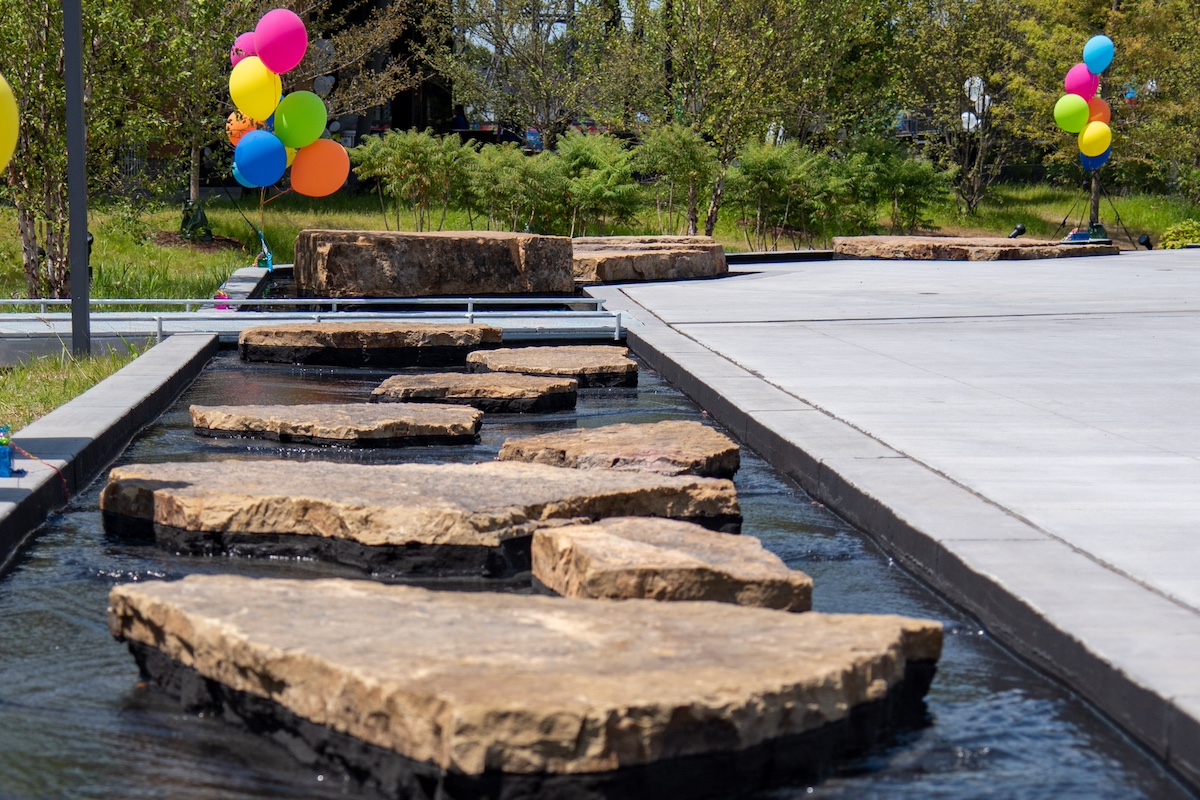 Stepping stones cross the Doermer Kids Canal at Promenade Park in Fort Wayne, Indiana, decorated with colorful balloons on a sunny day.