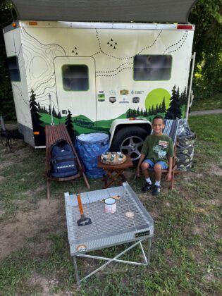 Child sitting by a camping trailer surrounded by outdoor gear and a Bronco-branded setup during Bronco Wild Fund Days, promoting outdoor exploration and family adventure.
