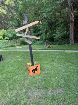 Wooden directional signpost with Bronco Wild Fund Days destinations in a grassy park, with a child playing cornhole in the background surrounded by nature.