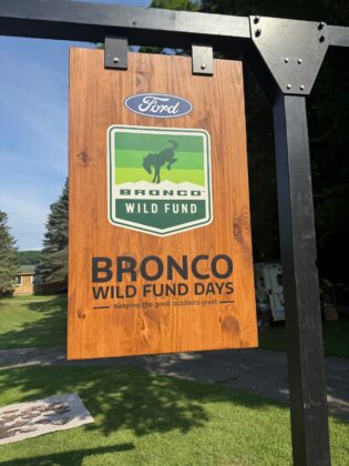 Wooden event sign for Bronco Wild Fund Days featuring the Ford and Bronco Wild Fund logos with the tagline 'Keeping the great outdoors great,' displayed in a scenic grassy area.