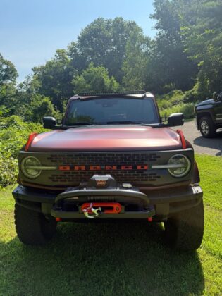 Front view of a red Ford Bronco with winch and off-road modifications displayed at Bronco Wild Fund Days, parked on grass near a wooded trail.