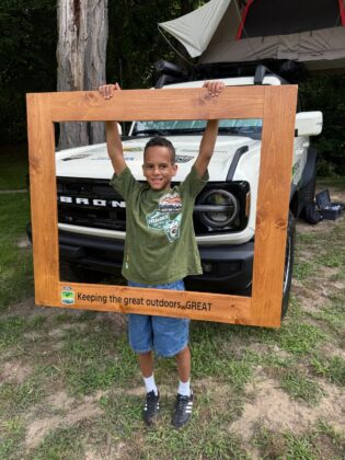 Smiling boy holding a wooden frame that says 'Keeping the great outdoors GREAT' in front of a rooftop tent-equipped Ford Bronco during Bronco Wild Fund Days.